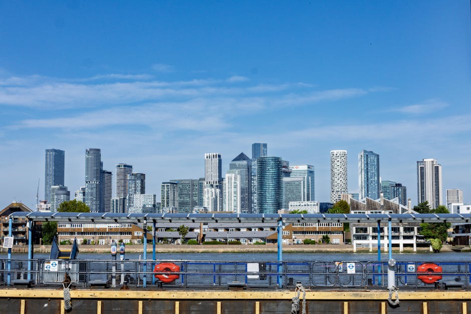 A cityscape featuring a modern glass skyscraper with reflective windows dominating the skyline, positioned centrally in the image. Surrounding it are other tall office buildings with sleek facades, so
