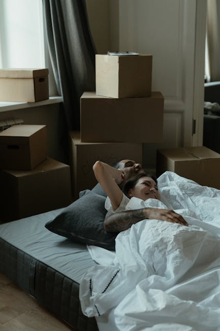 A sparsely furnished bedroom with white walls and a light wooden floor, featuring a large double bed with a simple wooden headboard covered by a plain white mattress. On either side of the bed are sma