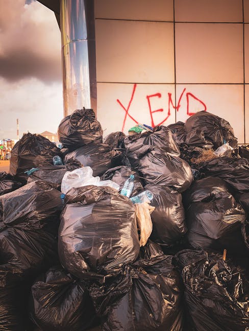 A large pile of black rubbish bags, some partially torn or bulging, is stacked against a modern, tiled building wall in an urban setting. The bags are filled with mixed waste, including visible plasti