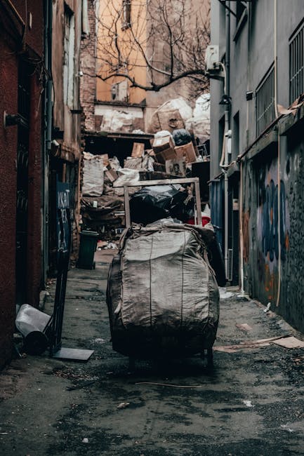 A large pile of black rubbish bags, some partially torn or bulging, is stacked against a modern, tiled building wall in an urban setting. The bags are filled with mixed waste, including visible plasti