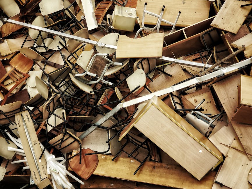 A large pile of discarded furniture and office equipment, including wooden desks, metal-framed chairs with plastic and wooden seats, and small storage units, all in various conditions, with some items